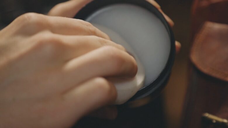A hand applies leather balm to a brown leather bag