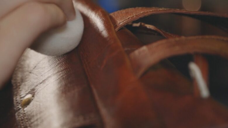 A hand wipes a brown leather bag with a soft cloth